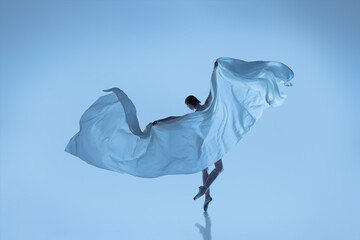 Portrait of flexible woman, graceful ballerina dancing with fabric, cloth isolated on blue studio...
