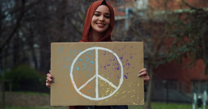 Cinematic shot of young pacifist Arabian muslim woman wearing hijab is showing placard with colorful peace symbol and smiling in camera during manifestation in green city park.