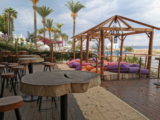 Cafe next to the beach. Tables and bar stools made from sawn wood, tents with soft, low mattresses, scattered multi-colored pillows and decorative lanterns