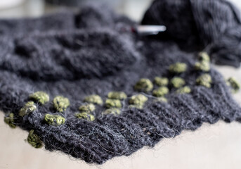 Green and black wool sweater in the process of being knitted. Photograph shows textured knitting including stripes and bobbles, knitting needles and balls of yarn.