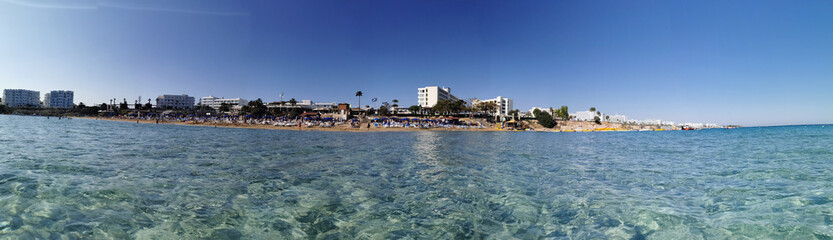 Protaras. Famagusta area. Cyprus. Panorama of Fig Tree Bay beach, people sunbathing and swimming, hotel buildings behind the beach against the blue sky. View from the sea.