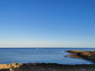 The rocky coast of the Mediterranean Sea from long-hardened lava, waves against a blue cloudless sky.