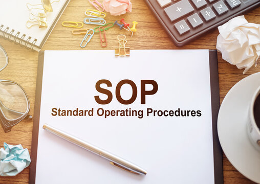 SOP acronym for Standard Operating Procedure, text on white paper next to office accessories on wooden desk.