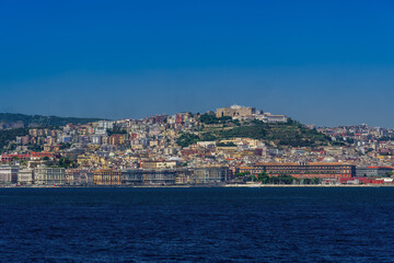 Naples, Italy city center coastal sea view under cloudless blue sky with Castel Sant'Elmo medieval fortress and waterfront buildings visible from a sailing ship on the gulf.