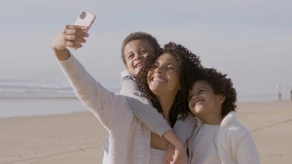 Happy mother and two children taking selfie while spending time at seashore. Smiling woman holding smartphone with stretched arm while family smiling at camera. Modern technology, happy family concept - Powered by Adobe