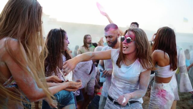 Joyful young woman in sunglasses smeared in gugal powder shares dry colors with friends at Holi festival in slow motion. Outdoor hindu holiday party. End of lockdown, covid pandemic, restrictions