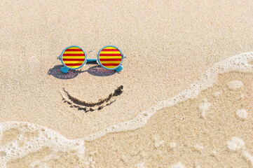 Sunglasses with flag of Catalonia on a sandy beach. Nearby is a sea lightning and a painted smile. The concept of a successful vacation in the resorts of Catalonia.