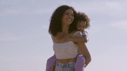 Beautiful young mother carrying young daughter on her back and laughing. Mother and child having fun outdoors on sunny day and playing. Parenthood, family concept