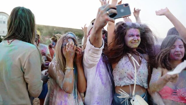 Cheerful people throw colorful powder, take selfie on smartphone and smile at Holi festival on beach in slow motion. Friends smeared in dry colors take group photo on phone. End of covid pandemic
