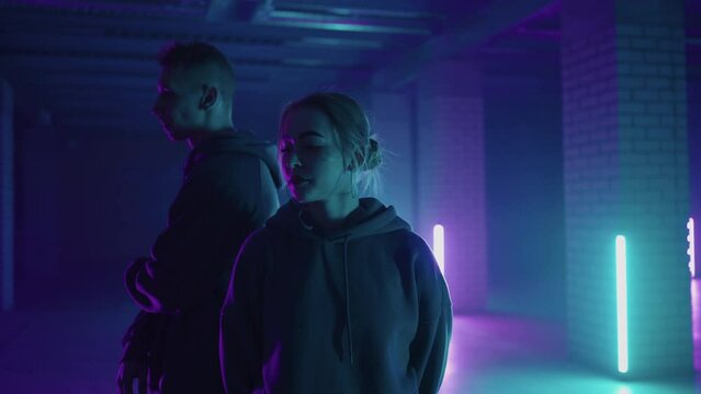 A duet of two professional dancers, a man and a woman. A stylish couple dances hip-hop freestyle together in synchronously in lard lit in blue purple hues with neon light