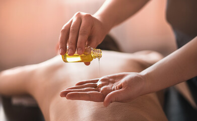 The masseur pours massage oil on his hand against the background of the back, a woman lying in the...