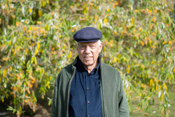 Portrait of aged man resting in spring park. Cheerful man in casual clothes and cap spending time outdoors standing on green trees background looking at camera. Leisure activity of aged people concept