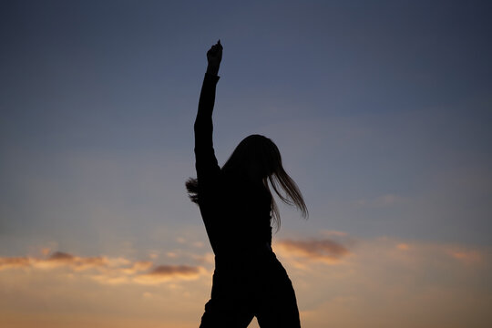 Dancing silhouette of attractive caucasian elegant girl at sunset or woman performing modern dance outdoor in sunny day against blue cloudy sky. Freedom, triumph or success concept. High quality image