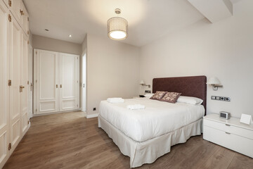 Bedroom with double bed with upholstered headboard, white bedside tables, fitted wardrobes with wooden doors and light wooden floors
