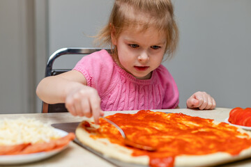 Small child cooking at home sitting at the table. Little girl making pizza in the kitchen. Kid learning to cook