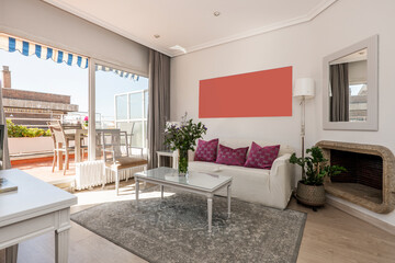 Living room with stone fireplace, sofa with white cover and purple cushions with large window leading to a sunny terrace