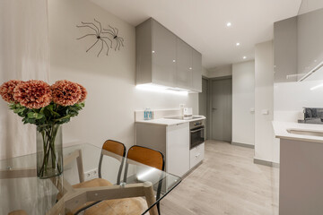 Glass dining table with floral centerpiece beside a kitchen with walls covered in glossy gray cabinets and white stone countertops
