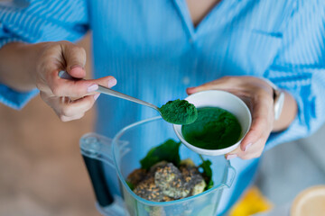 Close up woman adding spirulina green powder during making smoothie on the kitchen. Superfood...