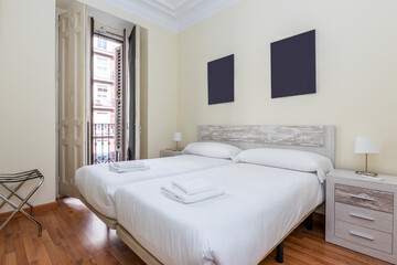 bedroom with two beds together with white bedding, wooden headboard and white lacquered carpentry, balcony with wooden shutters, matching bedside tables and wooden floors