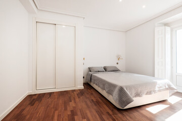 Fototapeta na wymiar Bedroom with a double bed with a gray bedspread, a built-in wardrobe with white sliding doors and a reddish oak parquet floor