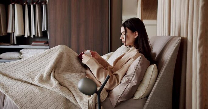 Young blonde girl is reading a book or a textbook. A beautiful woman is resting and relaxing at home in a cozy bedroom. Time for relax and education concepts. Cosy bed in a bedroom.