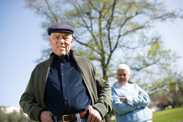 Portrait of aged couple posing for camera in park. Happy man in modern clothes and cap smiling looking at camera when woman standing behind him. Relations and leisure activity of aged people concept