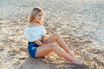 Young blonde woman in white top and jeans shorts sitting on the sand beach and enjoying the moment. Caucasian girl relaxing and enjoying peace on vacation. Selective focus, copy space