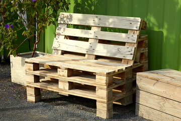 A wooden outdoor bench made of industrial pallets and a plant in a wooden box near a green fence....