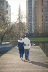 Back view of romantic aged couple in park. Hugging man and woman walking along wooden bridge over pond and looking at city and nature around. Aged couples relations and healthy lifestyle concept