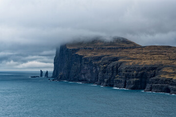 Steep cliff falling vertically into steel-blue ocean with two landmark rocks Risin and Kellingin, top covered with moody cold clouds