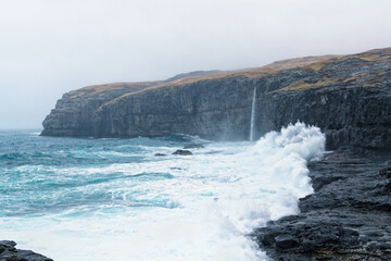 Ice-blue stormy sea breaking to rainy nordic black rocky beach with cliff and waterfall in background covered by misty clouds