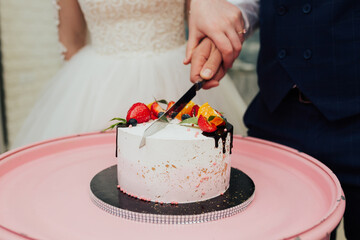 Bride and groom cutting stylish trendy wedding cake decorated with chocolate and berries at wedding reception in restaurant. Wedding couple holding knife and cutting together wedding cake 