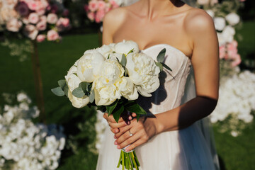 Elegant bride holding beautiful tender wedding bouquet of fresh peony and greenery in tender pastel white colours while standing near wedding arch in sunny day. 