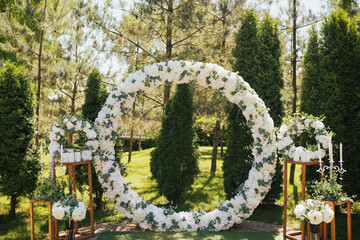 Wedding ceremony in the park in the sunny summer day. Beautiful wedding round arch decorated with white flowers, greenery and candles, outdoors.