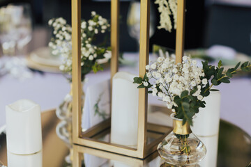 A branch of white flowers and greenery in a small vase, white candles on a table decorated for wedding ceremony.