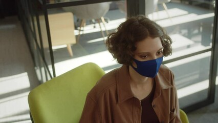 A young attractive girl-student is sitting in a blue protective face mask in the hall and looking at the camera, slow motion close up - Powered by Adobe