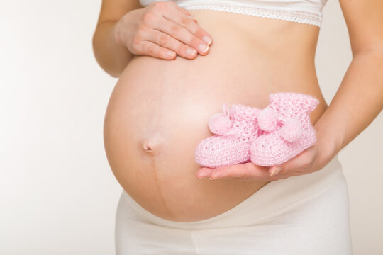 Young adult pregnant woman with big naked belly on light gray background. Hand holding and showing pink soft knitted socks for future newborn. Closeup. Preparing clothes for baby in pregnancy time.