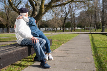 Fototapeta premium Modern senior couple kissing on bench in park. Grey-haired man and woman sitting on bench hugging and enjoying kisses on warm spring day. Relations, love and active rest of aged people concept