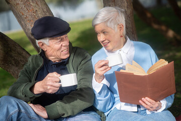 Close-up of senior couple sitting under tree. Caucasian man and woman sitting on ground, talking drinking tea and woman reading book with pleasure. Healthy lifestyle and rest of aged people concept