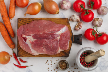 Organic raw pork meat on chopping board and fresh vegetables (tomatoes, onions, garlic, carrots, peppers) on Marble background, spices (coriander, pink and black pepper, chili) on the table.