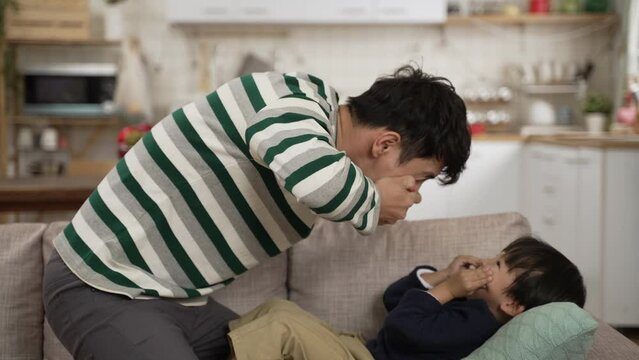 side view of excited Asian father enjoying playing with his baby son lying on the sofa. the man turns around and making funny silly face to make the boy laugh
