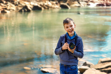 Photographer boy near a mountain river standing on the rock and looking at the camera. Kid enjoying the moment of the beautiful blue water in sunny day and smiling. Boy is holding photo camera.