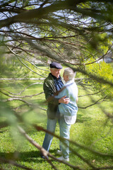 Loving aged couple spending day in spring park. Grey-haired man and woman standing on green grass hugging talking, looking at each other with love. Senior couples relations, love, active rest concept