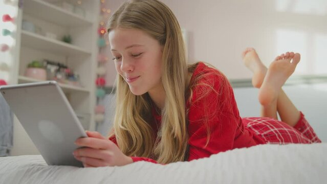 Teenage girl wearing orthodontic braces lying on bed at home streaming movie onto digital tablet and laughing - shot in slow motion