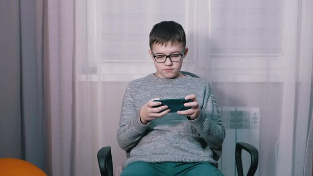 Surprised Child in Glasses Watching a Video in a Mobile App on a Smartphone. A teenager writes messages, SMS in a chat. Online learning, video game viewing at home. Internet addiction. Home interior.