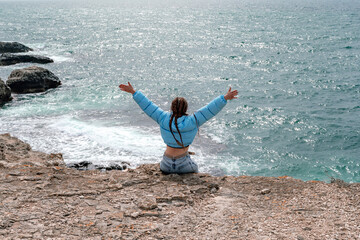 A woman in a blue jacket sits on a rock above a cliff above the sea, looking at the stormy ocean. Girl traveler rests, thinks, dreams, enjoys nature. Peace and calm landscape, windy weather.