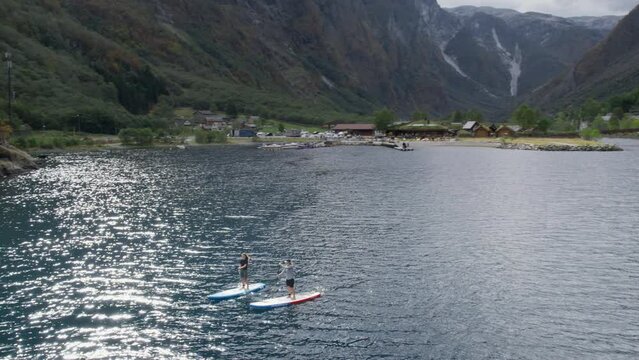 Young sporty energetic friends have fun stand up paddle boards in beautiful glacier fjord in western norway on warm weekend. Drone view on trendy lifestyle, bonding activity and tourism in Scandinavia