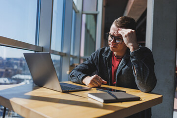 A freelancer in glasses looks into a laptop and he is angry while sitting at a table with a laptop and a notepad in a cafe during the day. Unlucky day. Working remotely from the office.