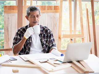 Fototapeta na wymiar Senior Asian man with a cup of hot drink looking at laptop computer while sitting at a table and resting in carpenter workshop. Home DIY in free time and hobby of older retired people.