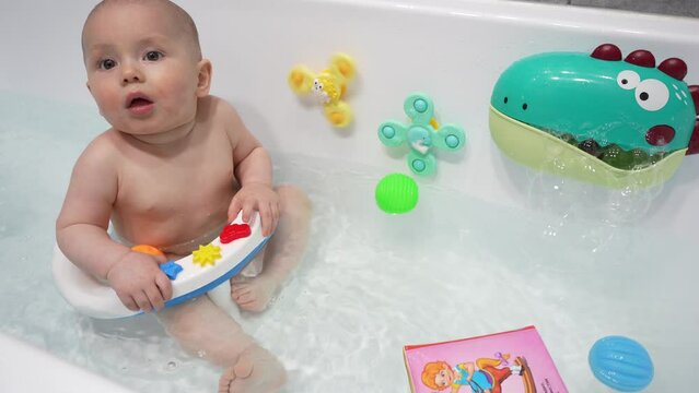 The baby boy bathes in the bathroom sitting in a chair. Happy toddler baby boy bathes in a white bath while sitting on a chair.